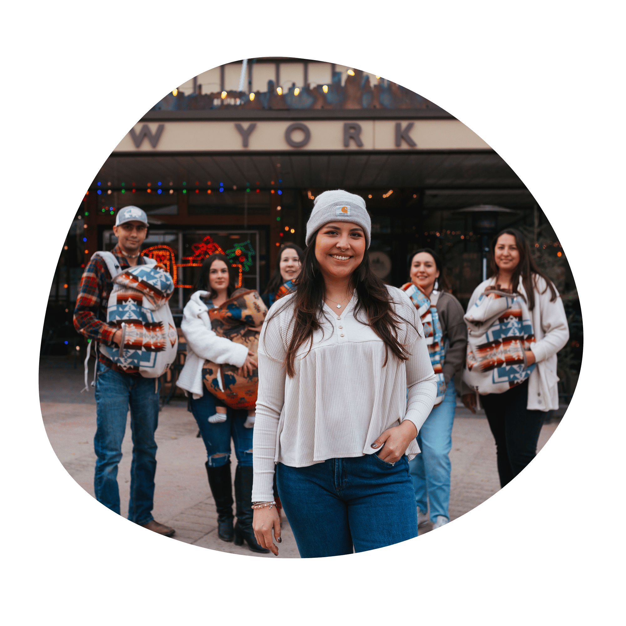 A line of babywearers stands in front of a store front. They all have moss bag covers over their baby carriers. The founder of Moss Bag Baby stands in front of the group smiling at the camera. 