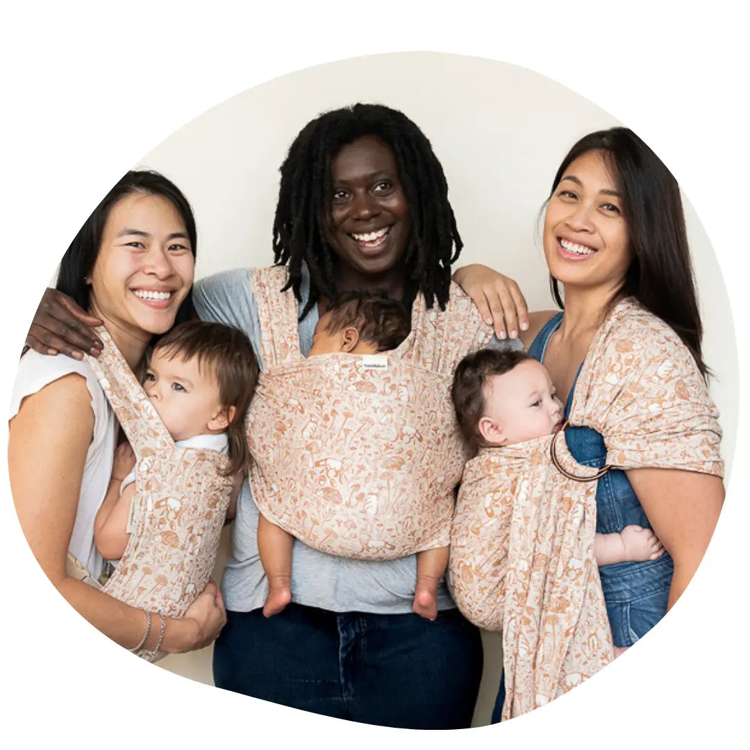 Three women stand with their arms around each other. All are wearing babies in different carriers.