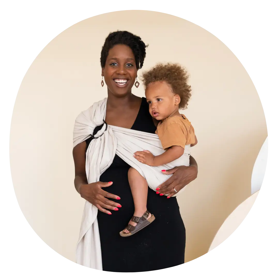 A Black woman stands in a clearing in the woods. She looks serenely down at her baby held in a floral ring sling