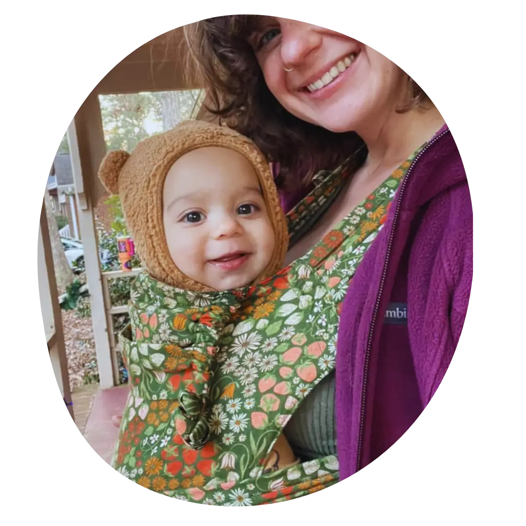 A woman in profile turns to smile at the camera while wearing a bundled and smiling baby in a front carry in a meh dai