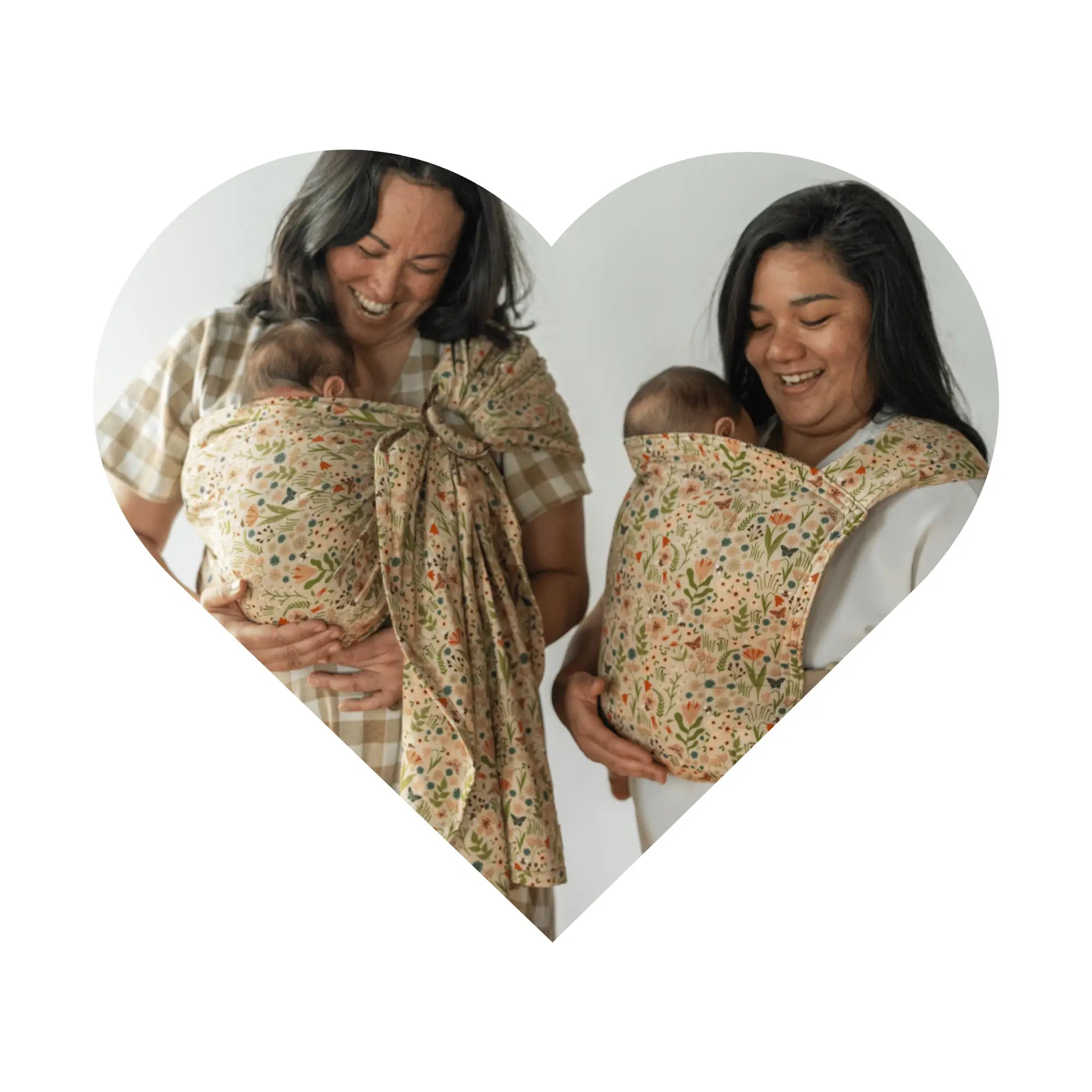 A heart frame of two woman smiling down at their babies worn in floral pattern carriers, one wrap and one Lark