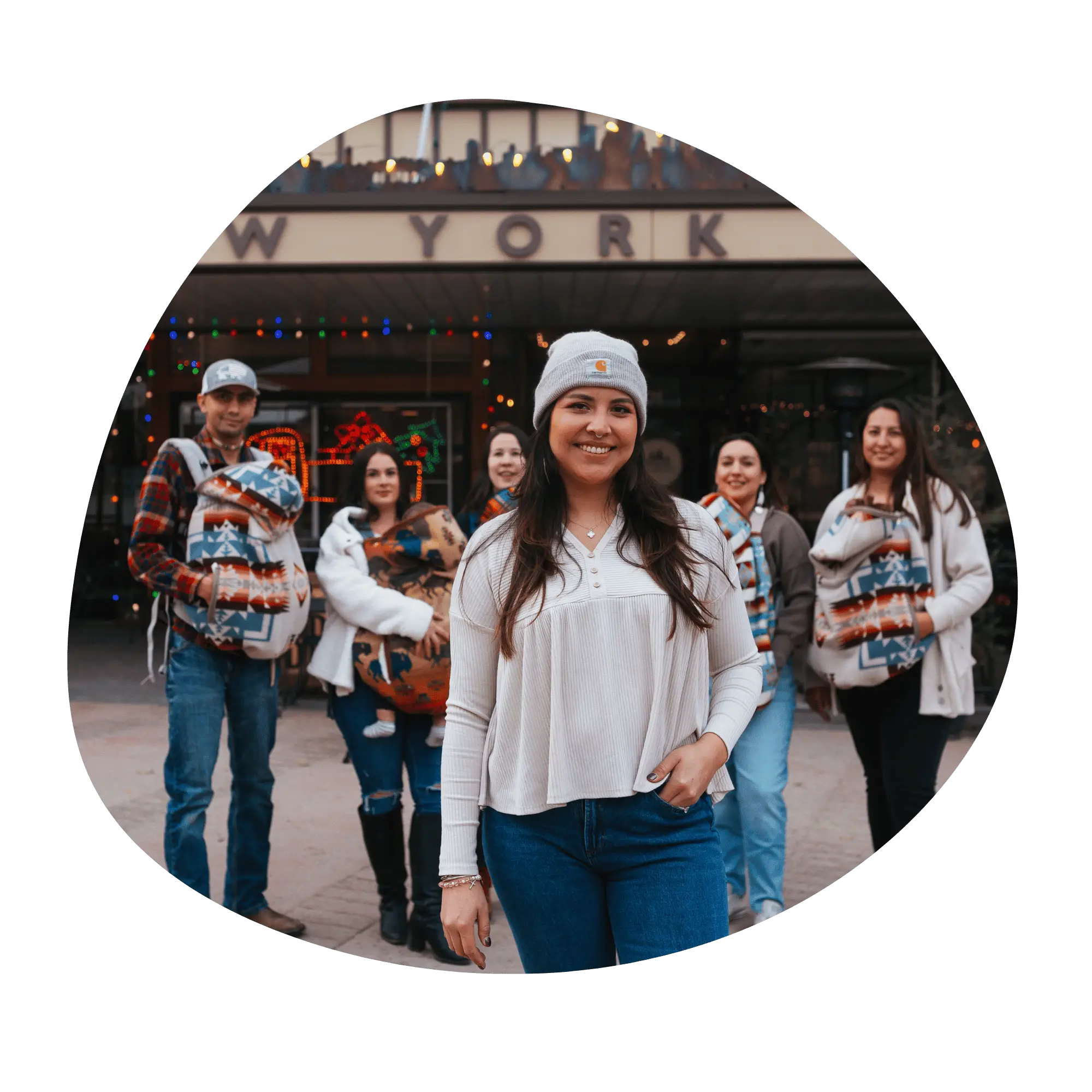 A line of babywearers stands in front of a store front. They all have moss bag covers over their baby carriers. The founder of Moss Bag Baby stands in front of the group smiling at the camera. 