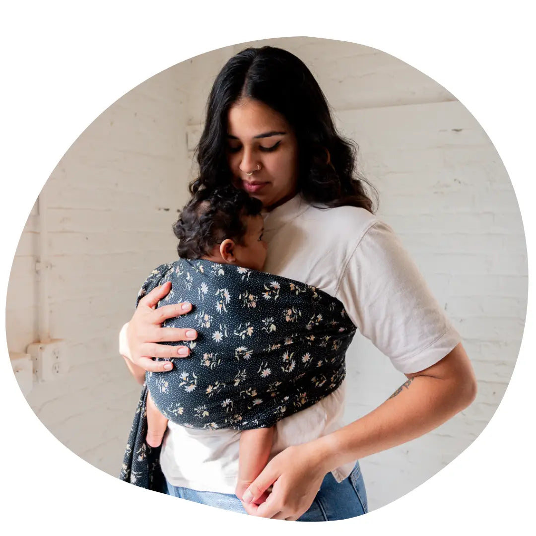 A woman stands while wearing an infant in a ring sling. She looks down at her baby.