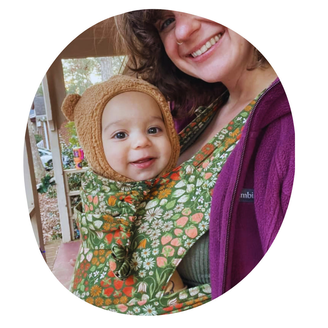 A woman in profile turns to smile at the camera while wearing a bundled and smiling baby in a front carry in a meh dai