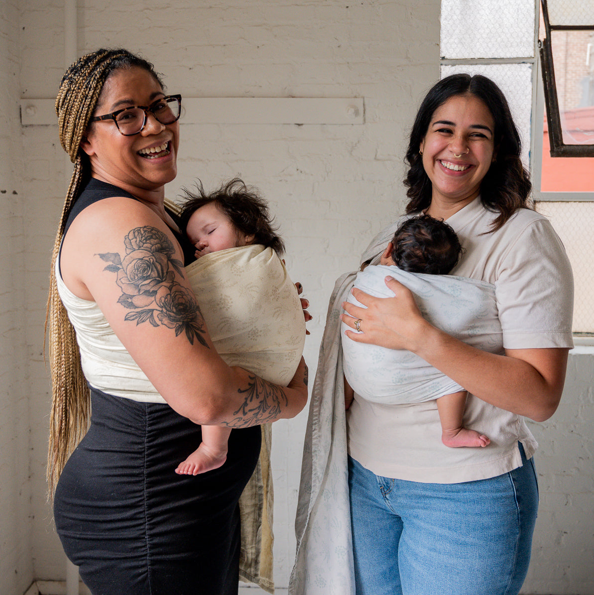 Two mothers, one on the left wearing a black dress and her baby in a cream ring sling and one on the right wearing jeans and her newborn in a light white ring sling carrier. 